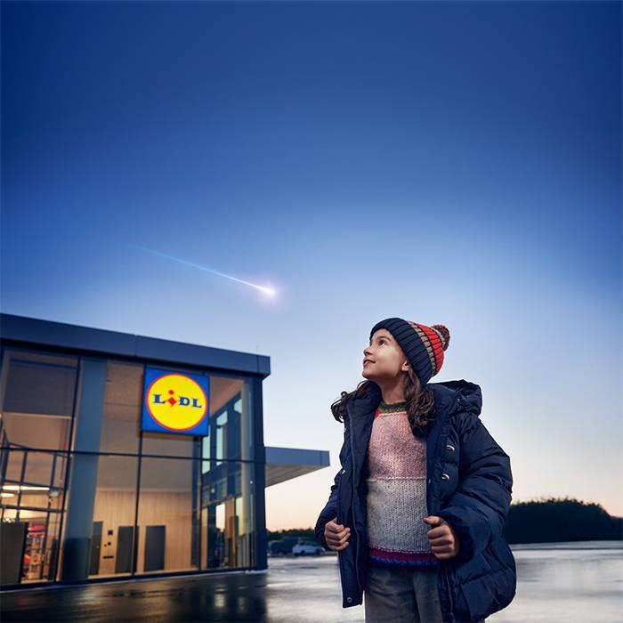 A child in winter clothing looks up at a shooting star above a Lidl store.