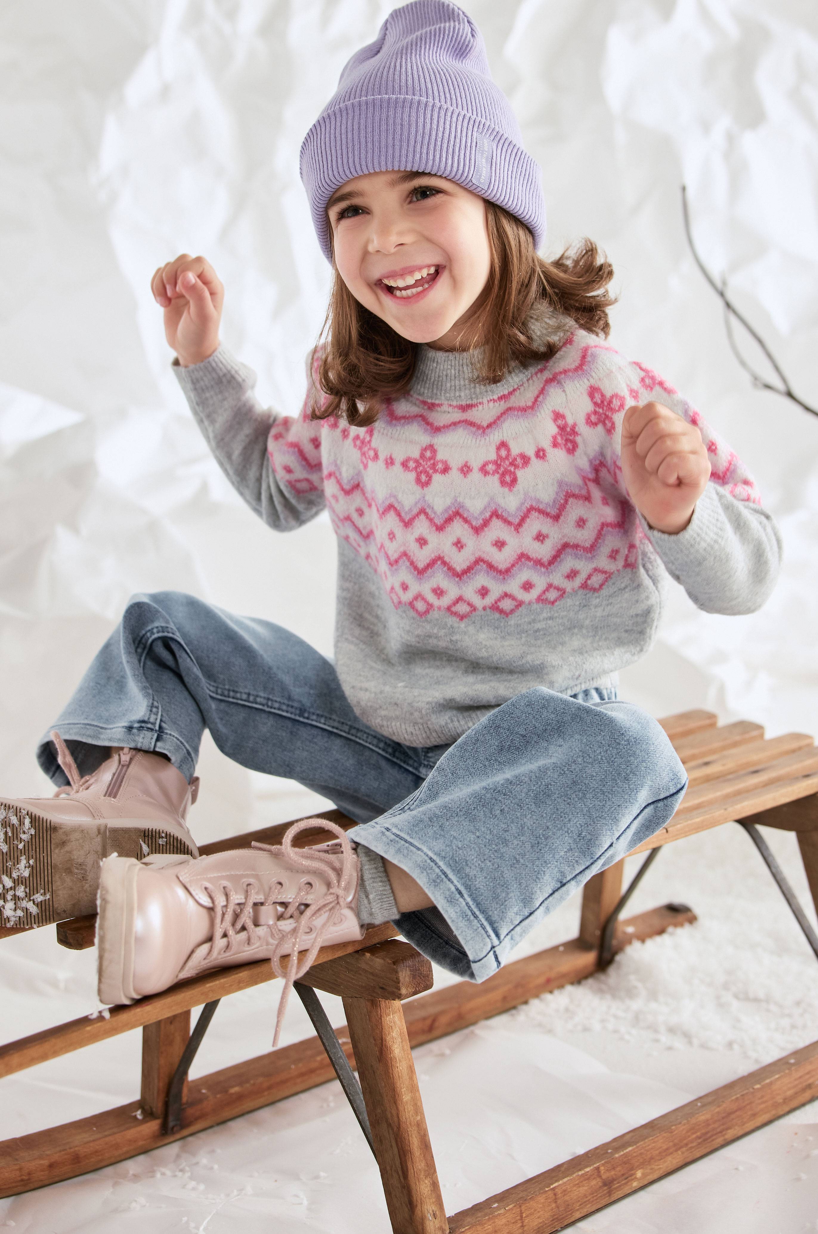 Girl in a purple beanie, knitted sweater with a Nordic pattern, and wide jeans, sitting on a wooden sled.