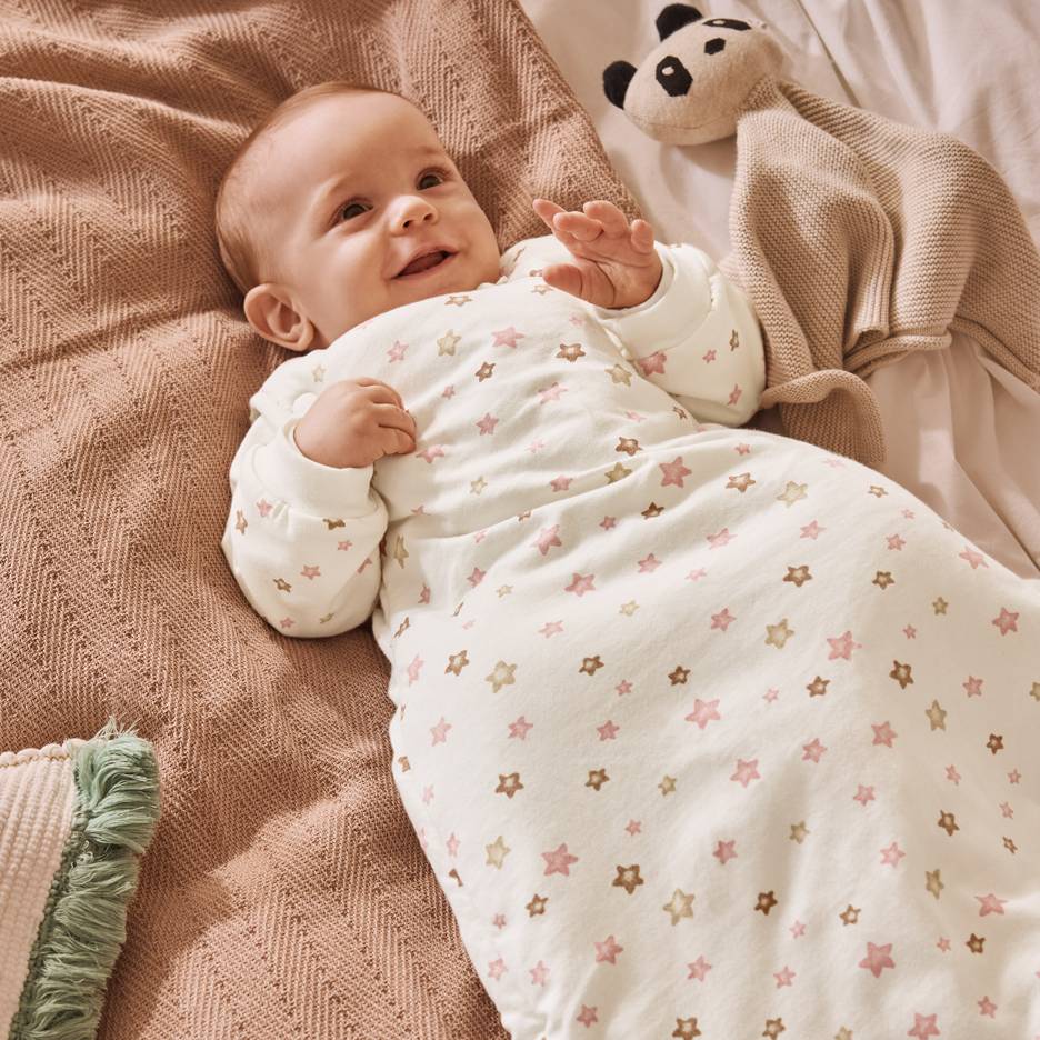 Smiling baby in a white sleeping bag with pink and brown stars, lying on a blanket.