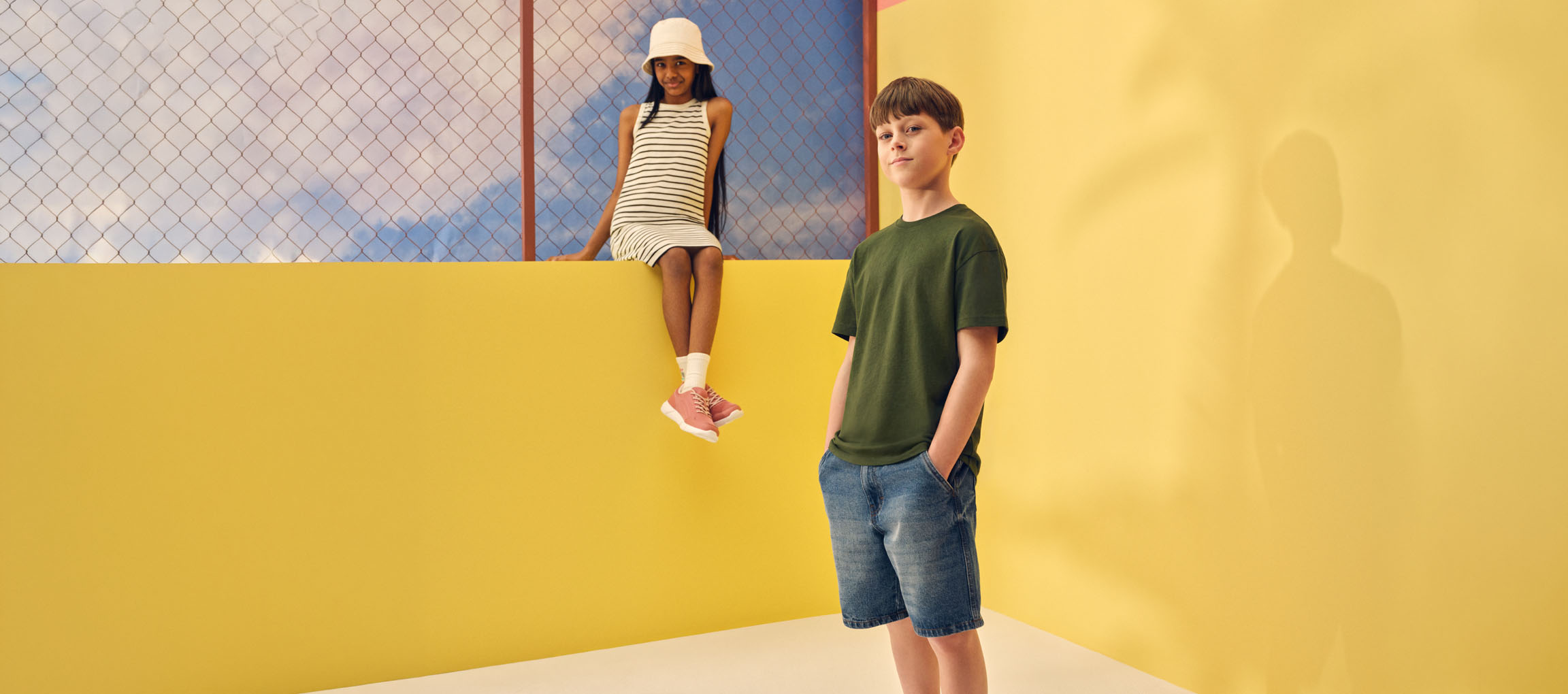 Two children in casual summer clothes: a girl in a striped dress and a boy in a green t-shirt and denim shorts.