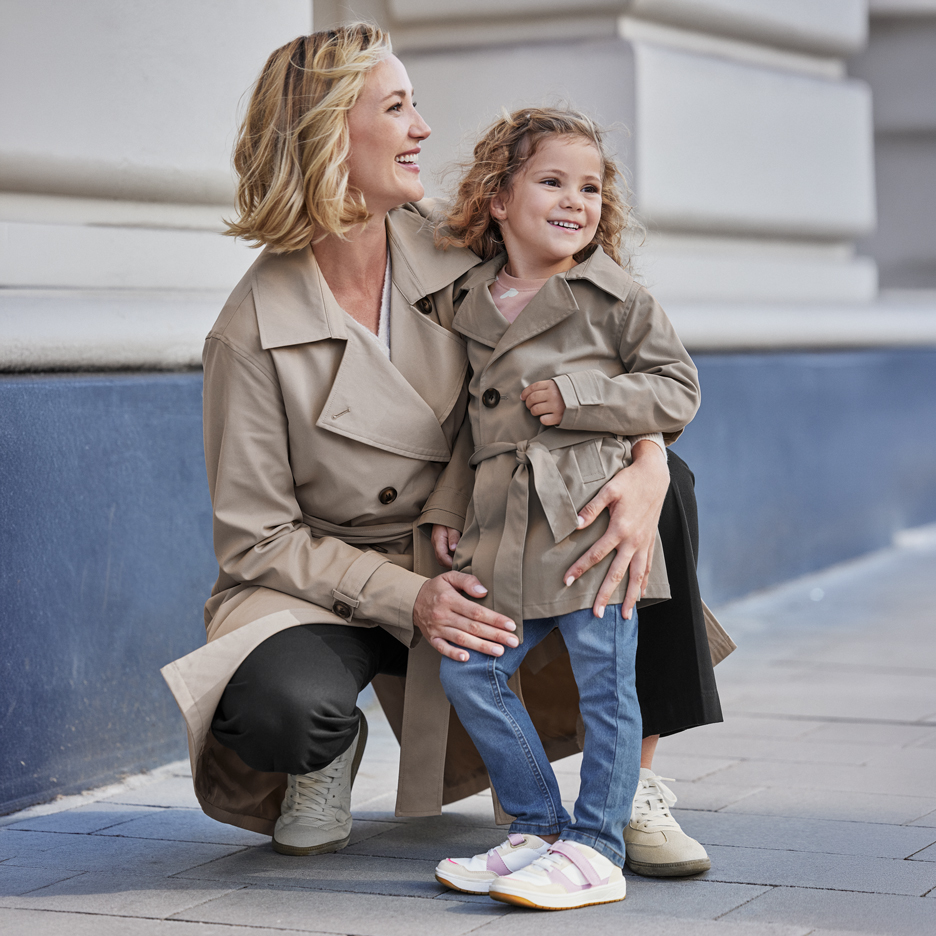 Two women in beige trench coats, an adult and a child, smiling outdoors.