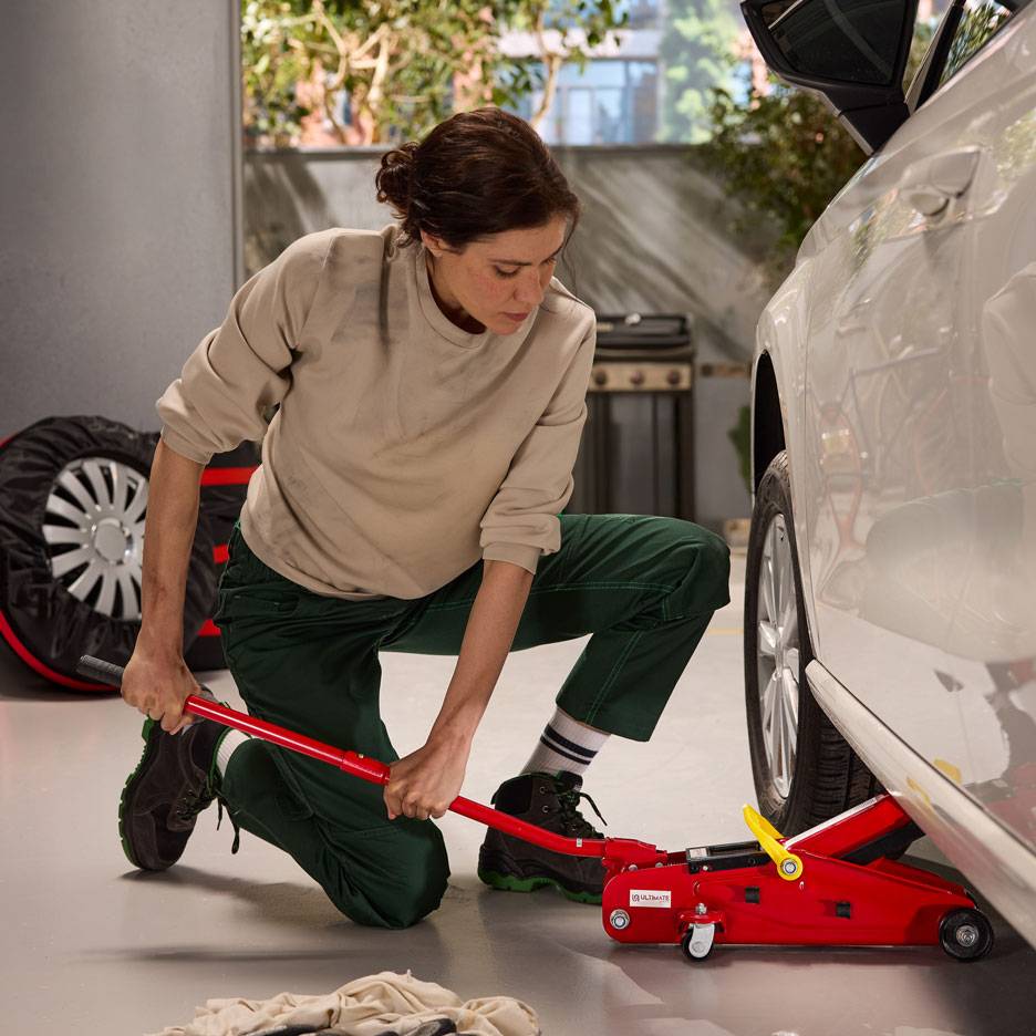 Woman using a red hydraulic jack to lift a white car, with a spare tire nearby.