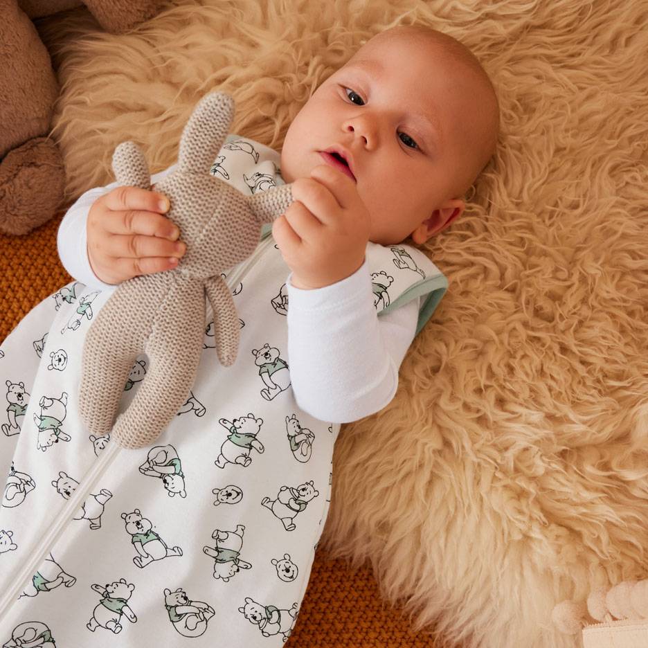Baby in white sleeping bag with bear pattern, holding a knitted bunny.