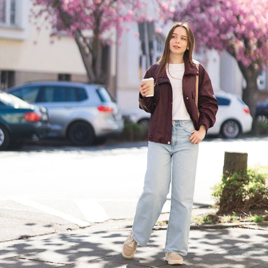 Woman in a jacket and jeans with coffee on a street with blooming trees.