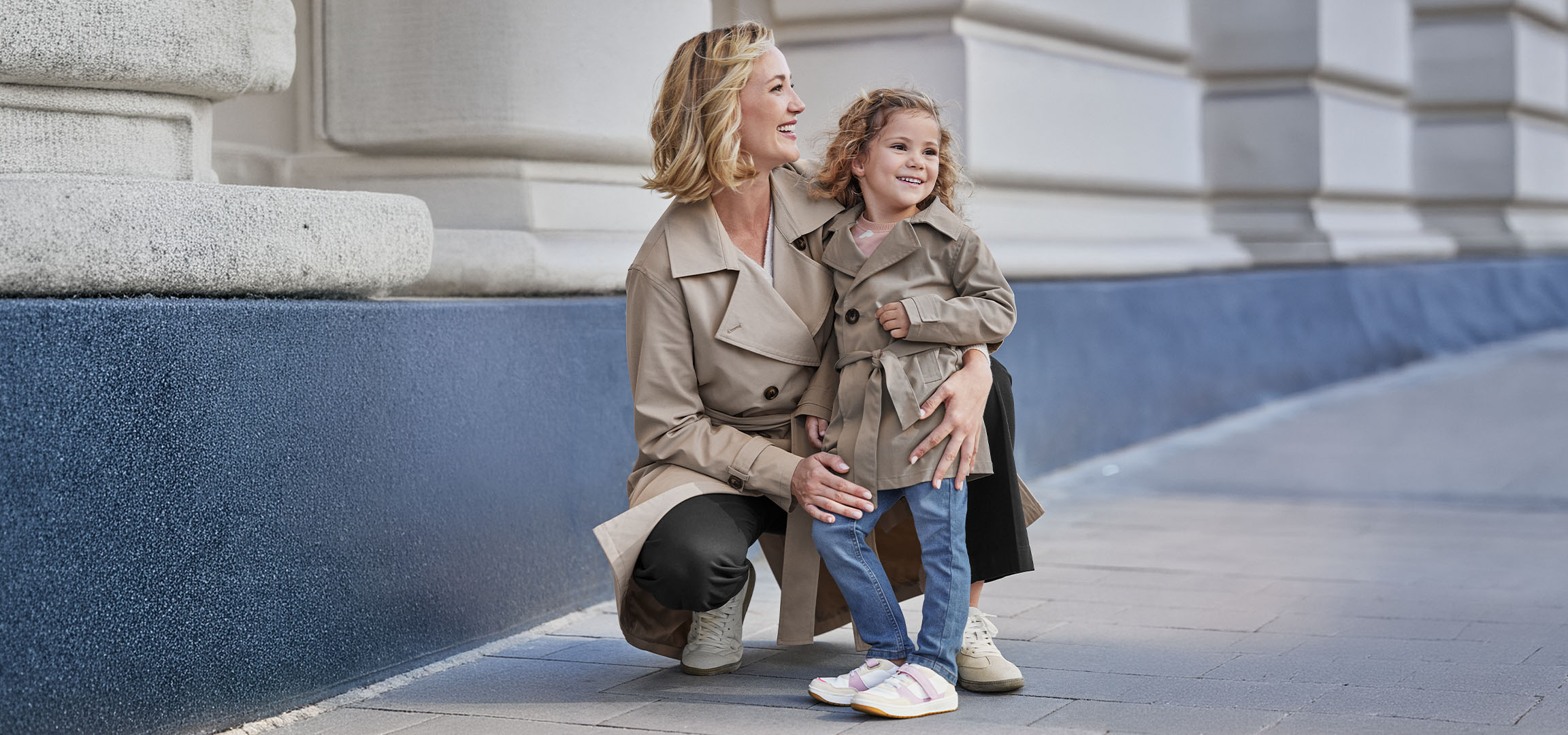 Two women in beige trench coats, an adult and a child, smiling outdoors.