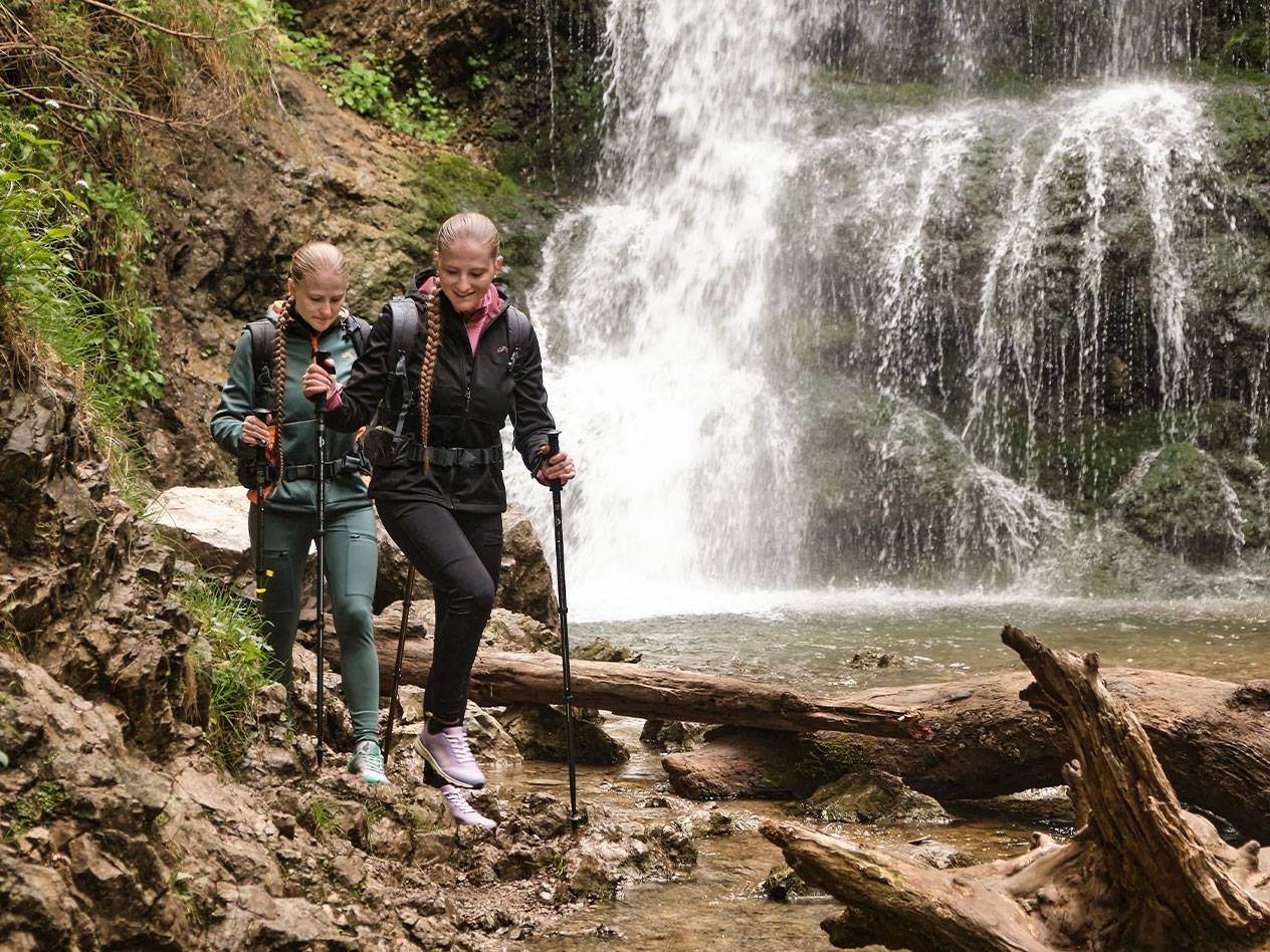 Two women with trekking poles hike a rocky path near a waterfall.