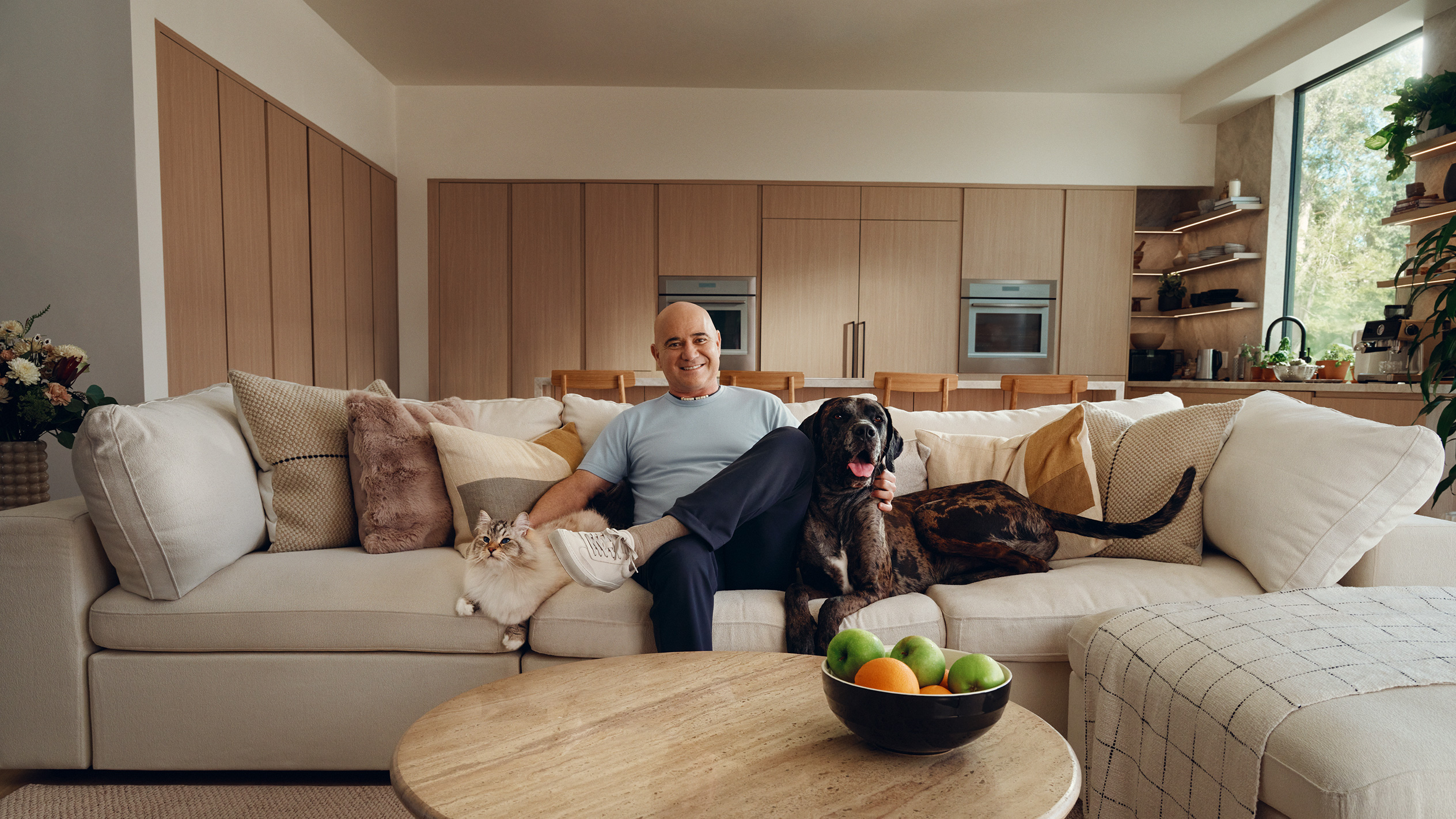 Man on sofa with cat and dog, fruit bowl on coffee table.
